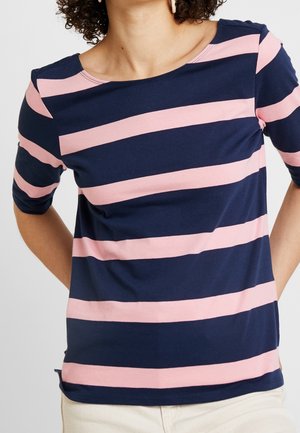 Kvinde iført en marineblå og lyserød horisontalt stribet skjorte med ærmer til albuerne, kombineret med lyse bukser.