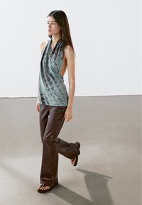 Een mouwloze teal top met een gedrapeerd ontwerp en een geweven textuur, gecombineerd met donkerbruine flare-pantalons en zwarte slippers.