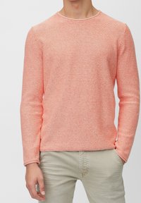 Korallfarbener, strukturierten Strickpullover mit langen Ärmeln und ribbeltem Halsauschnitt. Kombiniert mit hellbeigen Hosen, die Hände in den Taschen.