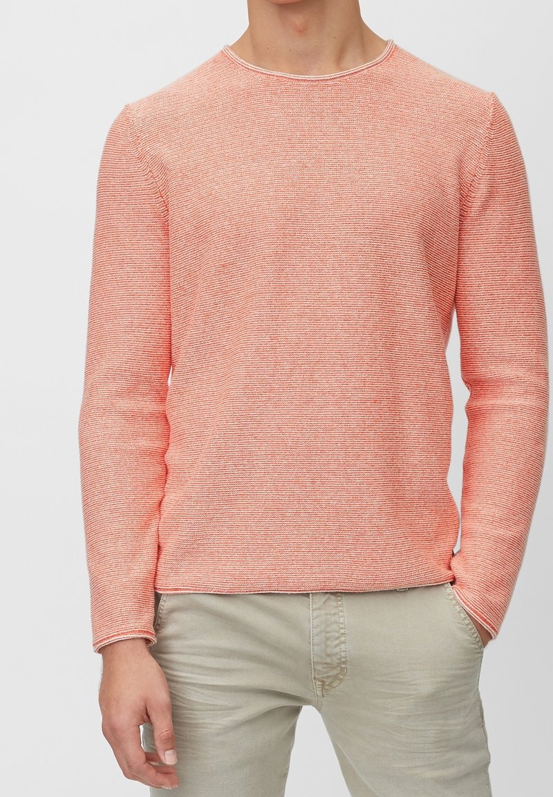 Korallfarbener, strukturierten Strickpullover mit langen Ärmeln und ribbeltem Halsauschnitt. Kombiniert mit hellbeigen Hosen, die Hände in den Taschen.