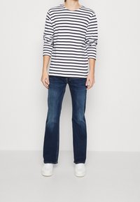 Camisa blanca de manga larga con rayas horizontales en azul marino, combinada con jeans azul oscuro y zapatillas blancas. Diseño simple con un ajuste relajado.