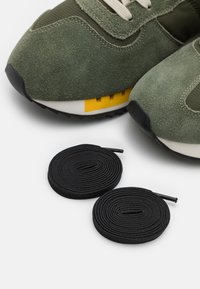 Baskets en daim vert avec des lacets beige et un accent jaune sur la semelle, associées à deux lacets ronds noirs, posées sur un fond blanc.