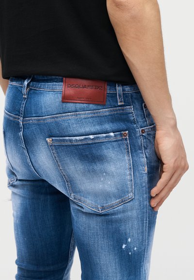 Κοντινό πλάνο ατόμου που φοράει μπλε τζιν με φθορές, με πίσω τσέπη και κόκκινη ετικέτα DSQUARED2, μαύρο πουκάμισο και εμφανές δεξί χέρι.