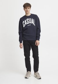Námornícka mikina s bielym nápisom "CASUAL", okrúhly výstrih, rebrované manžety, skombinovaná s čiernymi džínsami a béžovými teniskami s maskáčovými akcentmi.