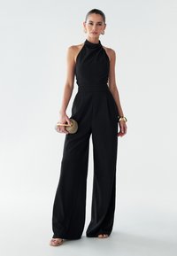 Melnā bezpiedurkņu kombinezona ar plašu starām. Iezīmēta ar teksturētu audumu, pieguļošu vidukli un elegantu siluetu. Aksesuārs - plecu soma.