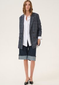 Blazer de cuadros gris de doble botonadura, camisa blanca de botones, pantalones cortos bermuda de mezclilla oscura con puños de mezclilla clara, mules negros de punta afilada.