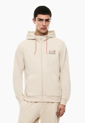 Junger Mann trägt einen beigen Kapuzen-Zip-Hoodie und passende Hose mit EA7 Emporio Armani-Logos auf Brust und Oberschenkel.