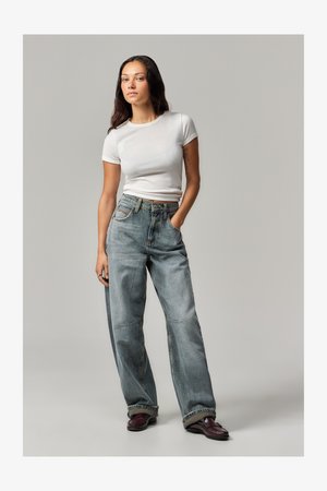 Jeans de ganga azul claro de perna larga com detalhes desbotados, cintura alta e bolsos frontais. Combinados com uma camisa justa de manga curta branca e sapatos bordôs.
