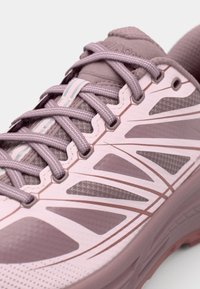 Hellrosa Sportschuh mit strukturiertem Obermaterial, reflektierenden Akzenten und einem gemusterten Design an den Seiten, abgerundet mit grauen Schnürsenkeln.