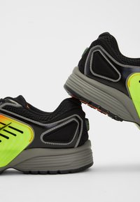 Sorte og grå sneakers med neongrønne accenter, mesh-overdel og en teksturert såle med dristig design og reflekterende detaljer.