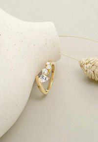 Anello d'oro con tre gemme trasparenti di forme diverse: una a goccia, una rotonda e una ovale, posizionato su uno sfondo di un vaso maculato.