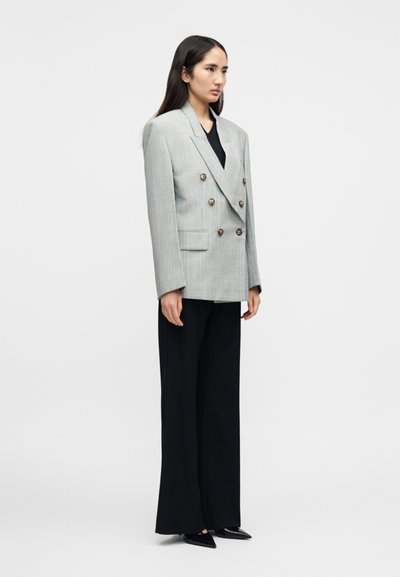 Δίπλο σακάκι σε ανοιχτό γκρι με μπλε ρίγες, με μεταλλικά κουμπιά και δομημένη σχεδίαση, συνδυασμένο με μαύρο παντελόνι φαρδιάς γραμμής.