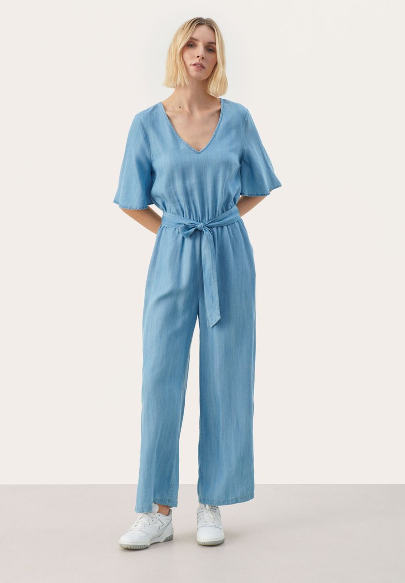 Jumpsuit azul claro de manga corta con un profundo escote en V, cintura ajustada con un nudo y diseño de pierna ancha, combinado con zapatillas blancas. Textura de tela suave.