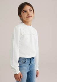 Maglietta bianca a maniche lunghe con texture arricciata e scollatura a volant, abbinata a jeans in denim blu, caratterizzati da una silhouette aderente e polsini arricciati.