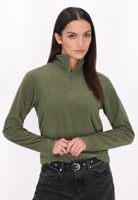 Pullover in pile verde oliva con colletto zip, maniche lunghe e texture liscia. Indossato con pantaloni neri a vita alta e una cintura.