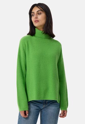 Grüner gerippter Rollkragenpullover aus weichem Strickmaterial mit langen Ärmeln und lockerer Passform. Getragen mit blauen Jeans.