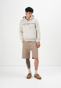 Jongeman in beige Tommy Hilfiger hoodie, tan shorts, beige sokken en bruine schoenen, staande met handen in zijn zakken, tegen een witte achtergrond.