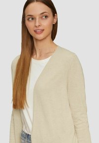 Beige cardigan met een V-halsontwerp, lichtgewicht stof en lange mouwen, gedragen over een wit shirt, gecombineerd met spijkerbroeken.