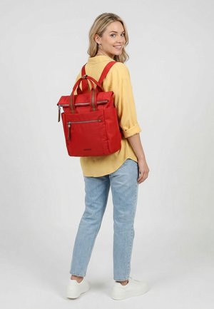 Nuori nainen, jolla on keltainen paita ja farkut, seisoo kyljellään hymyillen punainen reppu selässään yksiväristä taustaa vasten.