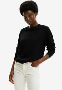 Maglione nero con un motivo testurizzato sulle maniche, scollo rotondo e polsini a costine. Abbinato a jeans aderenti di colore chiaro.
