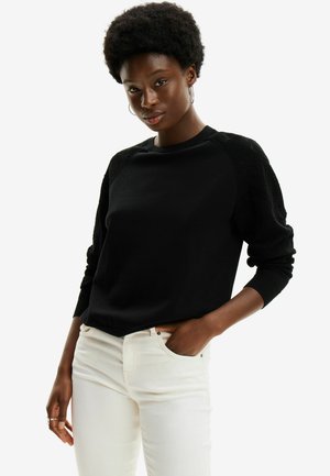 Maglione nero con un motivo testurizzato sulle maniche, scollo rotondo e polsini a costine. Abbinato a jeans aderenti di colore chiaro.