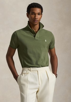 Nuori mies, jolla vihreä pikeepaita kohotetulla kauluksella ja luonnonvalkoiset laskostetut housut, kädet taskuissa, neutraali tausta.