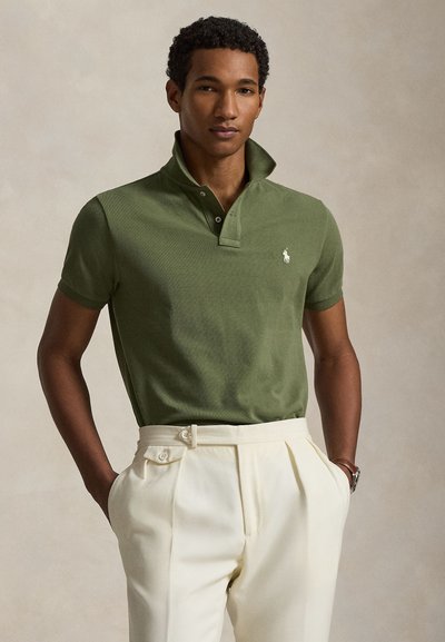 Giovane uomo che indossa una polo verde con colletto alzato e pantaloni plissettati color avorio, mani nelle tasche, sfondo neutro.