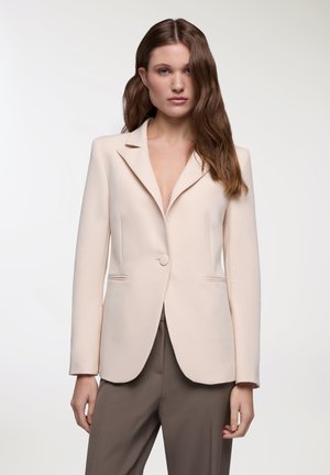 Blazer beige su misura con rever a lancetta, chiusura con un bottone e due tasche frontali. Tessuto liscio con un design minimalista.