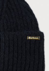 Gorro de malha preto com textura canelada e uma placa de logo dourada com "Barbour" na frente. Material de lã suave.