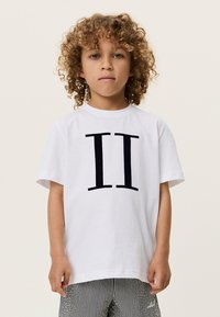 Λευκό βαμβακερό μπλουζάκι με μεγάλο μαύρο σχέδιο "II" στο μπροστινό μέρος. Κοντά μανίκια και στρογγυλή λαιμόκοψη. Ορατά ριγέ παντελόνια από κάτω.