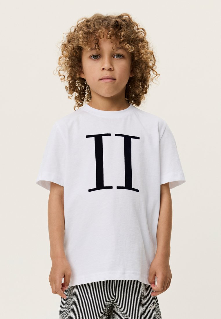 Λευκό βαμβακερό μπλουζάκι με μεγάλο μαύρο σχέδιο "II" στο μπροστινό μέρος. Κοντά μανίκια και στρογγυλή λαιμόκοψη. Ορατά ριγέ παντελόνια από κάτω.