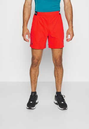 Mannelijk model draagt felrode sportshorts, een aansluitend teal T-shirt en zwarte hardloopschoenen, staand tegen een witte achtergrond.