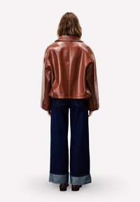 Persoon in een bruine leren jas en wijde donkerblauwe omgeslagen jeans, staande met zijn rug naar de camera op een witte achtergrond.