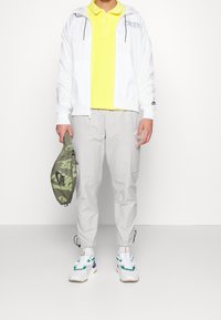 Bílá bunda na zip, žlutá košile s límečkem, světle šedé cargo kalhoty, zelená maskáčová taška a vícebarevné tenisky s masivními podrážkami.