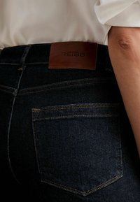 Mörka denimjeans med en läderbälte med märkningen "REISS". Har gyllene sömmar, bakfickor och en slim fit-design.