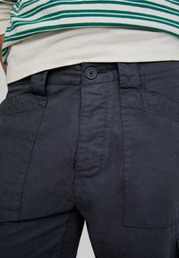Donkerblauwe cargobroek met een knoopsluiting, voorzien van voor- en achterzakken, textuurstof en zichtbare stikseldetails.