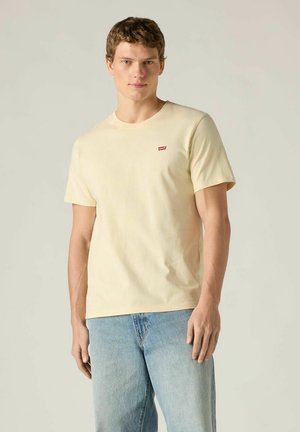 Camiseta de algodón amarillo claro con cuello redondo, mangas cortas y un pequeño logo rojo de Levi's en el pecho izquierdo, combinada con jeans azul claro.