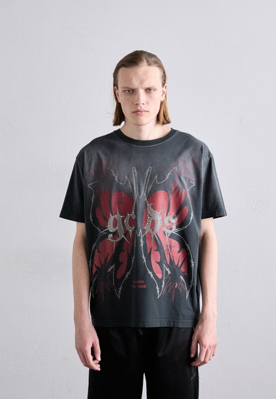 T-shirt oversize grigio scuro con una grafica di farfalla in rosso e argento, con elementi di filo spinato e testo in basso.