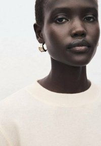 Gros plan d'une personne portant un pull texturé de couleur crème avec un col rond. Porte des boucles d'oreilles anneaux dorés, une peau lisse et des tons neutres.