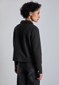 Veste noire courte avec un col, faite d'un tissu texturé, avec des poignets boutonnés, associée à un jean sombre. Vue de dos.