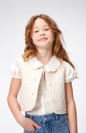 Beige quiltet vest med blomsterdesign, avrundede kanter og knapper, brukt over en hvit bluse med Peter Pan-krage. Blå denimshorts.