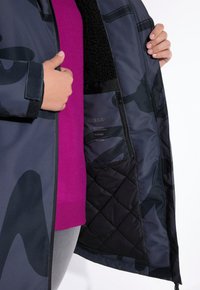 Cappotto impermeabile di un blu scuro con motivo mimetico, foderato in pelliccia nera e tasca interna con zip. Maglione magenta visibile sotto.