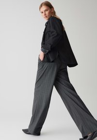 Un blazer noir surdimensionné avec des poches sur un pantalon large rayé gris, complété par un tissu lisse et des chaussures noires à talons pointus.
