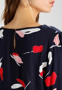 Granatowa bluzka z kwiecistym wzorem w kolorze czerwonym i różowym, z detalem w postaci dziurki z tyłu oraz miękkiego, teksturowanego materiału. Krótkie rękawy i luźny krój.