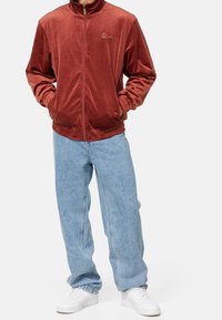 Röd sammetsjacka med dragkedja, hög krage, elastiska muddar och resår i midjan. Kombinerad med ljusblå vida jeans och vita sneakers.