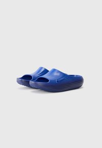 Une paire de sandales bleu Nike à enfiler, avec un bout ouvert et des semelles texturées sur un fond gris clair uni.