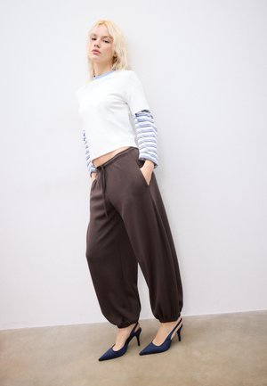 Giovane donna con capelli biondi che indossa una maglia a maniche lunghe a strati bianca e a righe, pantaloni marroni larghi e scarpe con tacco slingback blu navy, davanti a una parete semplice.