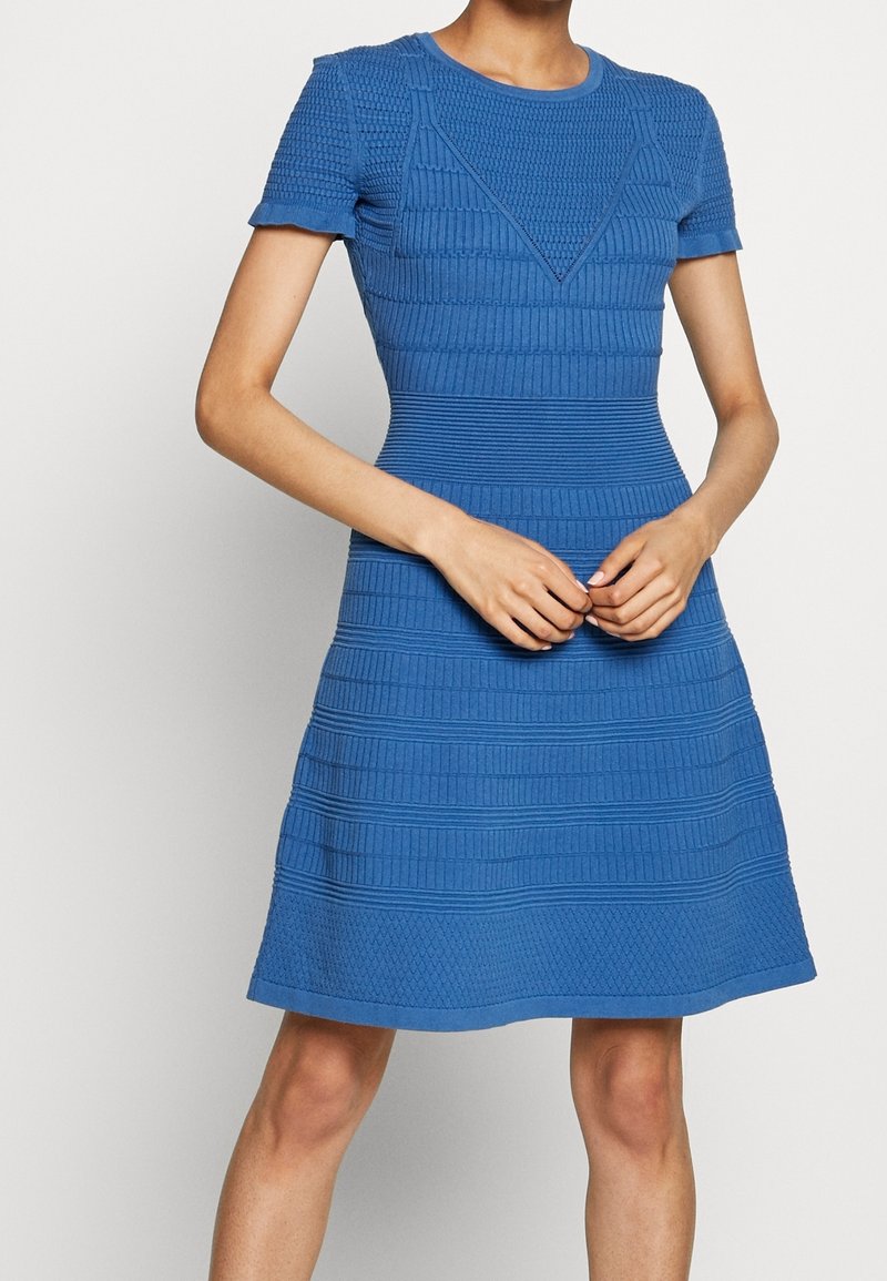 Femme portant une robe en maille texturée bleue à manches courtes avec des motifs horizontaux et géométriques, debout les mains jointes devant elle.