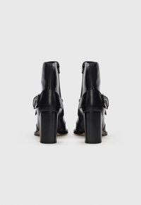 Par de botines de cuero negro con tacones cuadrados, correas con hebillas y cremalleras traseras sobre un fondo blanco.
