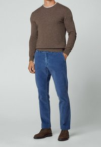 Homme portant un pull marron, un pantalon en velours côtelé bleu, et des bottes en daim marron, debout avec une main dans la poche sur un fond uni.